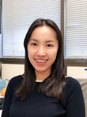 A young asian woman with mid-length brown hair wears a navy blue sweater and smiles at the camera in an office.